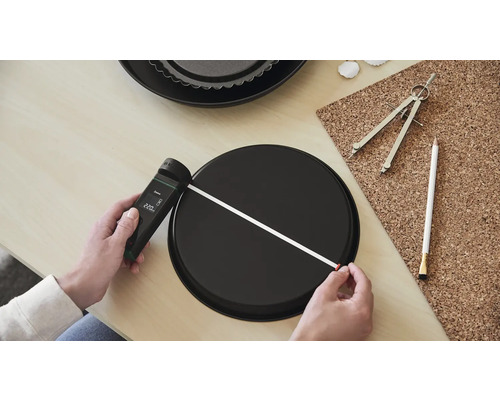 Eine Person misst mit einem Laser-Entfernungsmesser den Durchmesser einer runden Backform. Eine zweite Backform, ein Zirkel und ein Stift liegen auf dem Tisch.