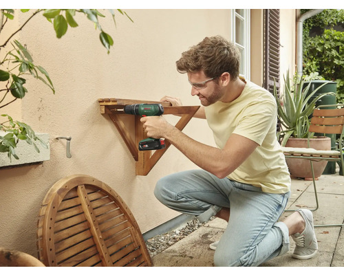 Mann montiert mit einem Akku-Bohrschrauber einen Holztisch an einer Wand im Freien.