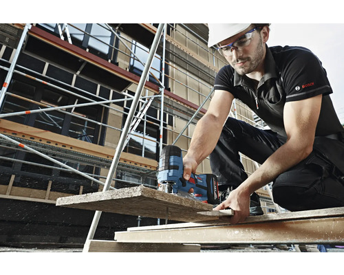Ein Bauarbeiter mit Helm und Schutzbrille sägt mit einer Stichsäge ein Holzbrett auf einer Baustelle. Im Hintergrund ist ein Gerüst zu sehen. Bosch Logo.