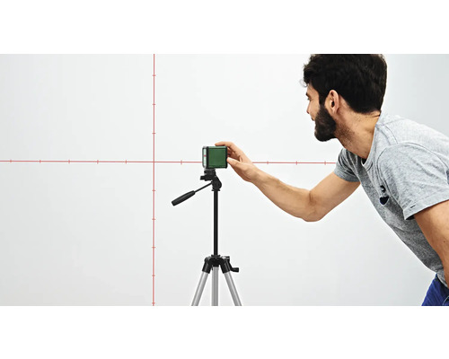 Ein Mann justiert einen Linienlaser auf einem Stativ, der rote Linien an eine Wand projiziert.