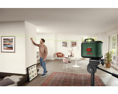 Bosch Logo. Ein Mann markiert eine grüne Laserlinie mit einem Bleistift an einer Zimmerwand. Der grüne Linienlaser steht auf einem Stativ.