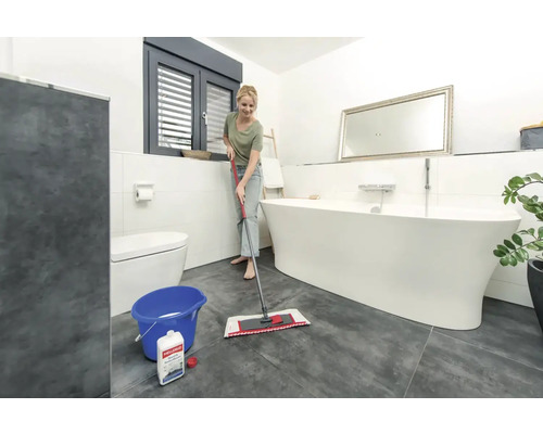 Helles Badezimmer mit freistehender Badewanne, Toilette und Frau, die mit einem Mopp und Eimer den Boden wischt.