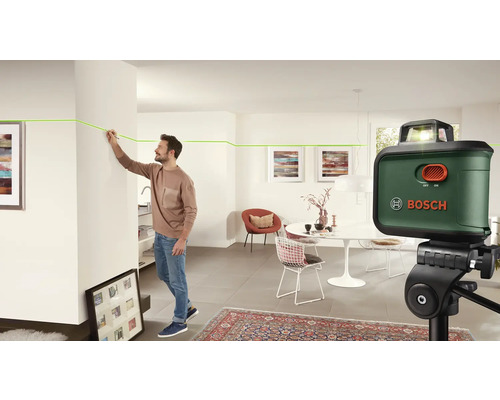 Ein Mann benutzt einen Linienlaser von Bosch in einem Wohnzimmer, um eine Linie an einer Wand zu markieren. Der Laser steht auf einem Stativ.