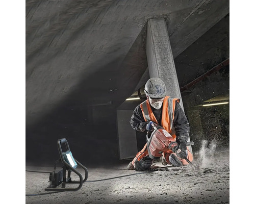 Ein Bauarbeiter benutzt einen Bohrhammer auf einem Betonboden. Ein Scheinwerfer leuchtet die Baustelle aus.