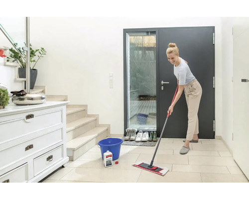 Frau wischt Fliesenboden mit einem Flächenwischer, Reinigungsmittel und Eimer im Flur eines Hauses.