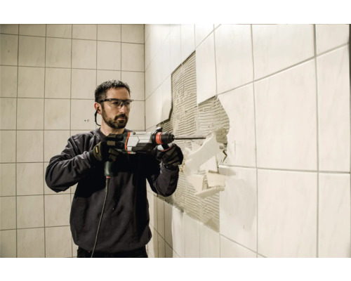 Ein Mann mit Schutzbrille und Handschuhen entfernt Fliesen mit einem Bohrhammer von einer Wand.