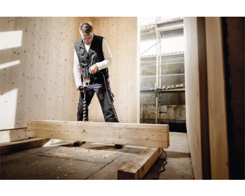 Handwerker bohrt mit einer Bohrmaschine in ein Holzbrett in einem Raum mit Holzpaneelen.
