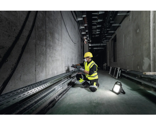 Ein Arbeiter mit Schutzhelm und Schutzweste bohrt mit einer Bohrmaschine in eine Betonwand, beleuchtet von einem Arbeitsstrahler.
