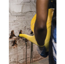 Eine Person gießt mit einer Gießkanne Wasser in eine beschädigte Ziegelwand, um diese vor der Reparatur zu befeuchten.