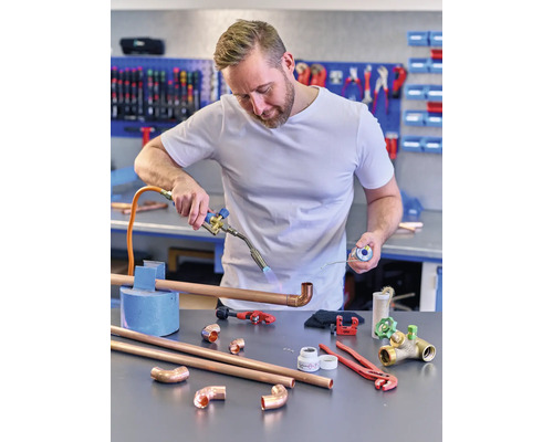 Ein Mann lötet ein Kupferrohr mit einem Schweißbrenner, während er Lötzinn hält. Verschiedene Werkzeuge und Kupferrohrfittings liegen auf dem Tisch.