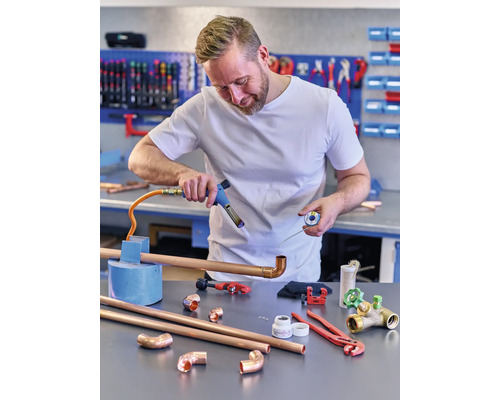 Ein Mann lötet Kupferrohre mit einem Lötkolben in einer Werkstatt. Verschiedene Werkzeuge und Rohre liegen auf dem Tisch.