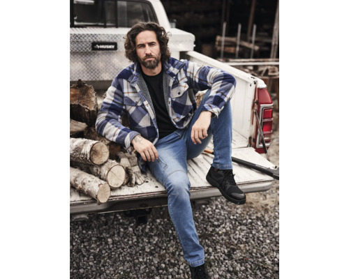 Mann sitzt auf der Ladefläche eines Pickups mit Holzscheiten, gekleidet in Jeans, Hemd und Sicherheitsschuhen.