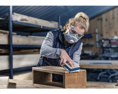 Frau schleift Holzbox mit Schleifklotz. Sie trägt Schutzbrille und Atemschutzmaske.
