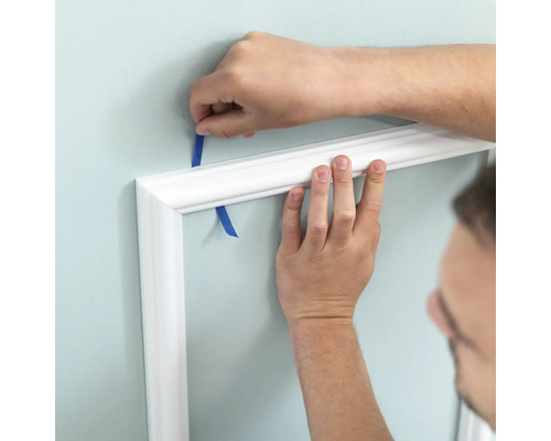 Person befestigt weiße Zierleiste mit Klebeband an der Wand.