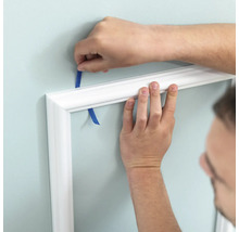 Person befestigt eine weiße Zierleiste mit Klebeband an der Wand.