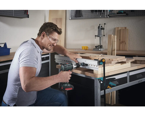 Mann bohrt mit einer Bohrmaschine durch eine Holzplatte mit Bohrschablone auf einer Werkbank.