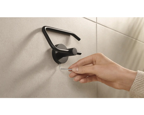 Eine Hand montiert einen schwarzen Toilettenpapierhalter mit einem Inbusschlüssel an einer Wand.