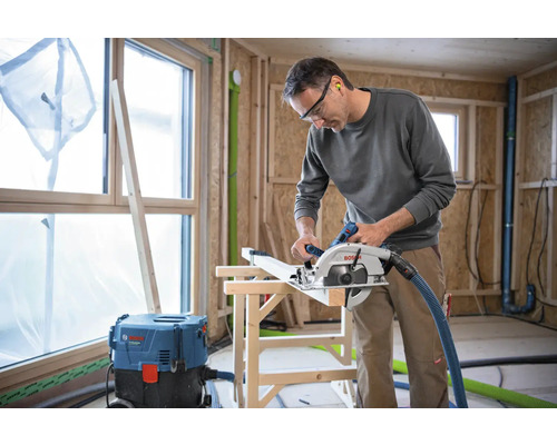 Ein Mann sägt Holz mit einer Handkreissäge in einer Baustelle. Im Hintergrund sind ein Fenster und ein Staubsauger zu sehen.