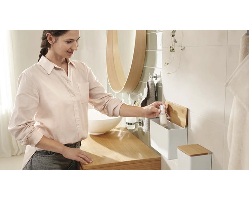 Helles Badezimmer mit Waschbecken, Spiegel und einer Frau, die eine Flasche aus einem Wandregal nimmt.