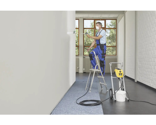 Ein Handwerker mit Atemschutzmaske streicht eine Wand mit einem Farbsprühgerät von einer Leiter aus.