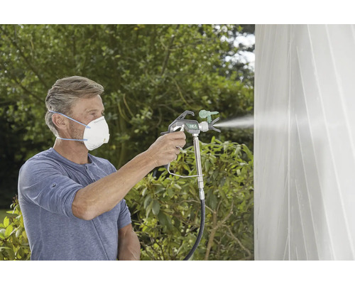 Mann mit Atemschutzmaske lackiert eine Wand mit einem Farbsprühsystem im Außenbereich.