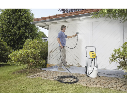 Eine Person streicht mit einem Farbsprühgerät ein Gebäude im Freien. Das Sprühgerät steht auf einem Rollwagen neben einem Eimer.