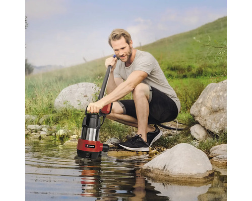 Mann hockt am Ufer eines Gewässers und hält eine Schmutzwasserpumpe.