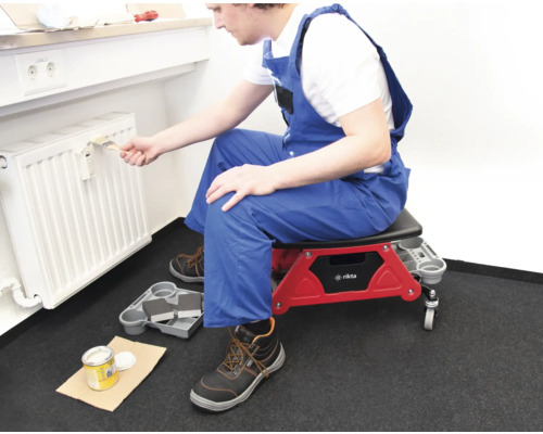 Eine Person sitzt auf einem Rollhocker und streicht einen Heizkörper mit einem Pinsel.