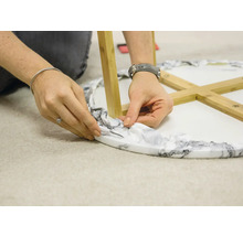 Eine Person schneidet überschüssiges Verpackungsmaterial von einem Tisch mit Marmoroptik und Holzbeinen ab.