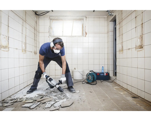 Mann mit Schutzbrille, Gehörschutz und Mundschutz arbeitet mit einem Abbruchhammer in einem gefliesten Raum.