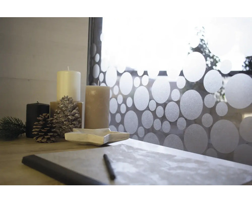 Dekorative Anordnung mit Kerzen, Tannenzapfen, sternförmiger Schale und Schreibmaterial vor einem Fenster mit Punktmuster.