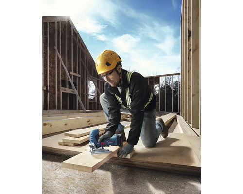 Ein Handwerker mit Schutzhelm und Sicherheitsbrille sägt ein Holzbrett mit einer Stichsäge auf einer Baustelle.