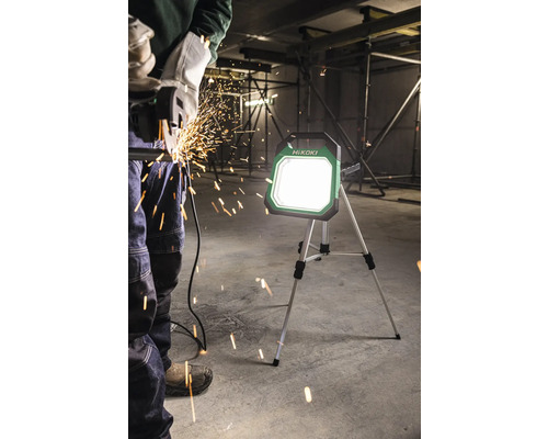 HiKoki Logo. Eine Person schneidet Metall mit einer Flex, während ein HiKoki Baustrahler die Baustelle beleuchtet.