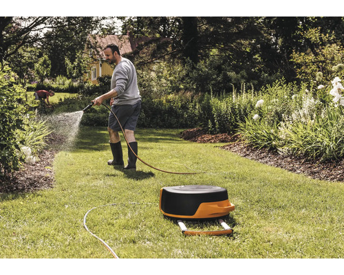 Ein Mann bewässert mit einem Gartenschlauch einen Rasen neben einer Schlauchtrommel.