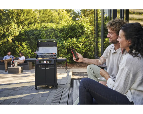 Szene mit einem Holzkohlegrill, zwei Personen im Vordergrund und zwei Personen auf einer Couch im Hintergrund.