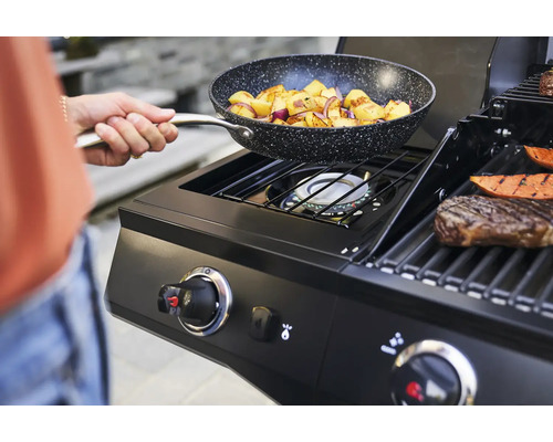 Person hält eine Bratpfanne mit Bratkartoffeln über einem Gasgrill.