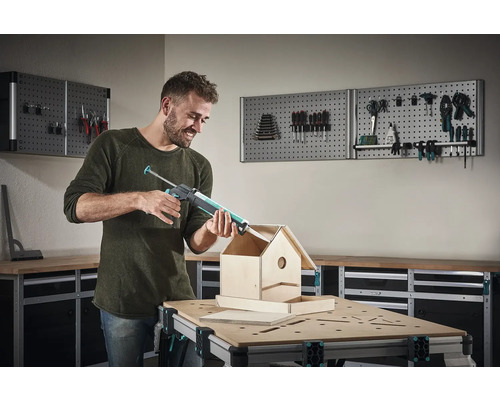 Mann verwendet eine Kartuschenpistole zum Bau eines Vogelhauses an einer Werkbank vor einer Werkzeugwand.