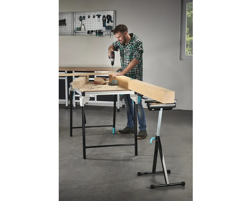 Mann bohrt mit einem Akkubohrer in ein Holzstück auf einem Arbeitstisch, im Hintergrund Werkzeuge an einer Werkzeugwand