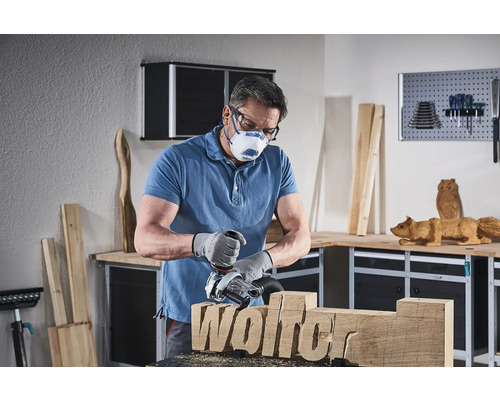 Mann arbeitet mit einer Holzfräse an einem Holzstück mit dem Schriftzug Wolfer, trägt dabei eine Schutzmaske und Schutzbrille in einer Werkstatt.