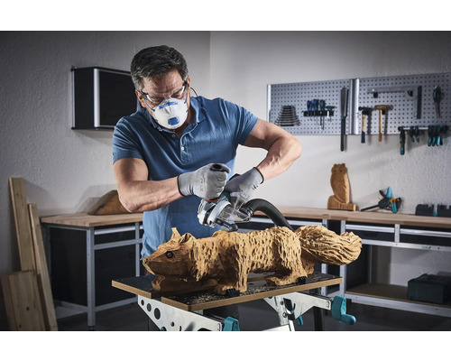 Mann schleift mit einer Schleifmaschine eine Eichhörnchen Holzfigur. Er trägt eine Maske, eine Schutzbrille und Handschuhe.