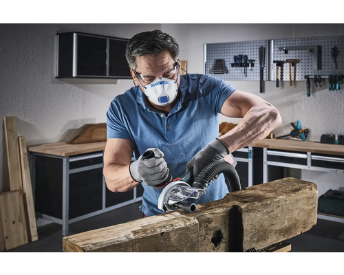 Ein Mann schleift ein Holzstück mit einem Elektrowerkzeug in einer Werkstatt, wobei er eine Schutzmaske und Handschuhe trägt.