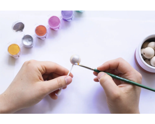 Eine Person bemalt eine Holzkugel mit Farben und einem Pinsel. Farbtöpfe und weitere Holzkugeln sind im Bild zu sehen.