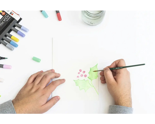 Person malt mit Aquarellfarben Stechpalmenblätter auf Papier. Aquarellstifte liegen daneben.