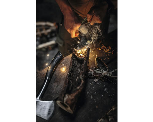 Ein Mann entzündet mit einem Feuerstarter ein Feuer in einem Holzscheit, neben dem eine Axt liegt.