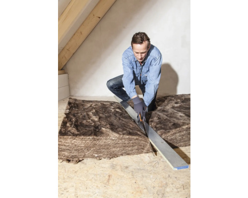 Mann schneidet Dämmmaterial auf einem Dachboden mit einem Messer und einem Richtscheit zu.