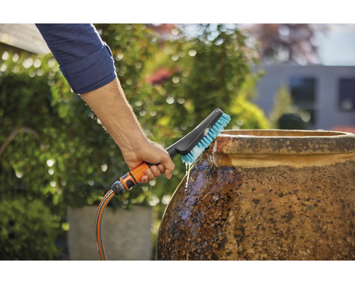 Eine Person reinigt einen Topf mit einer Bürste, die an einen Wasserschlauch angeschlossen ist, im Garten.