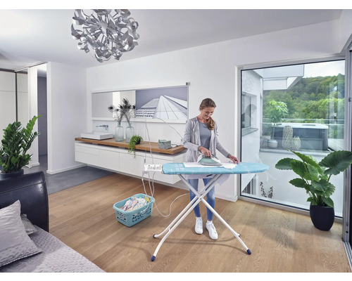 Frau bügelt Wäsche auf einem Bügeltisch in einem hellen Raum.