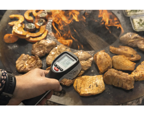 Eine Person misst die Temperatur von Speisen mit einem Infrarotthermometer über einem offenen Feuer.