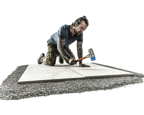 Ein Mann kniet und verlegt Terrassenplatten mit einem Gummihammer auf einem Kiesbett.