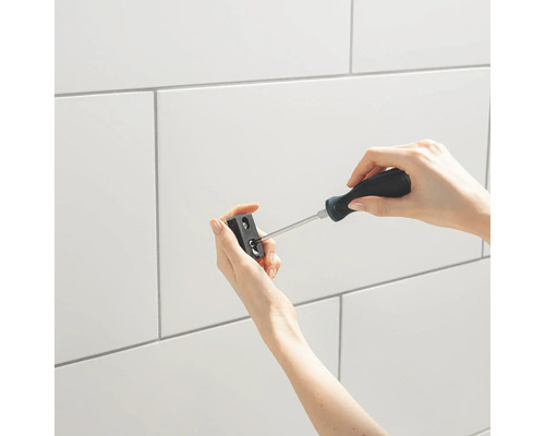 Eine Person befestigt eine Halterung mit einem Schraubenzieher vor einer Wand aus weißen Fliesen.
